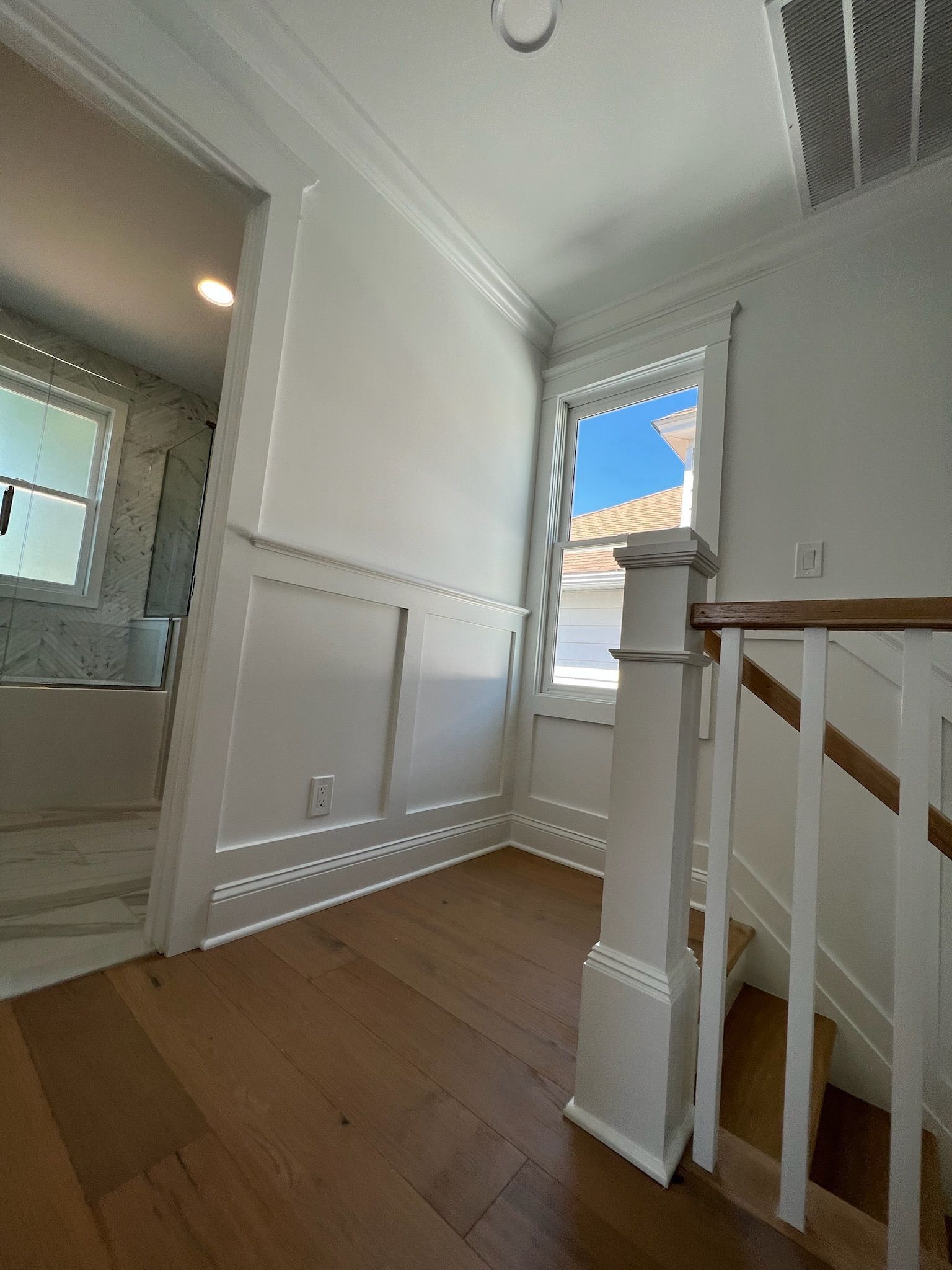 A staircase landing with white wainscoting, wooden floors, and a railing, looking toward a bright window and a doorway.