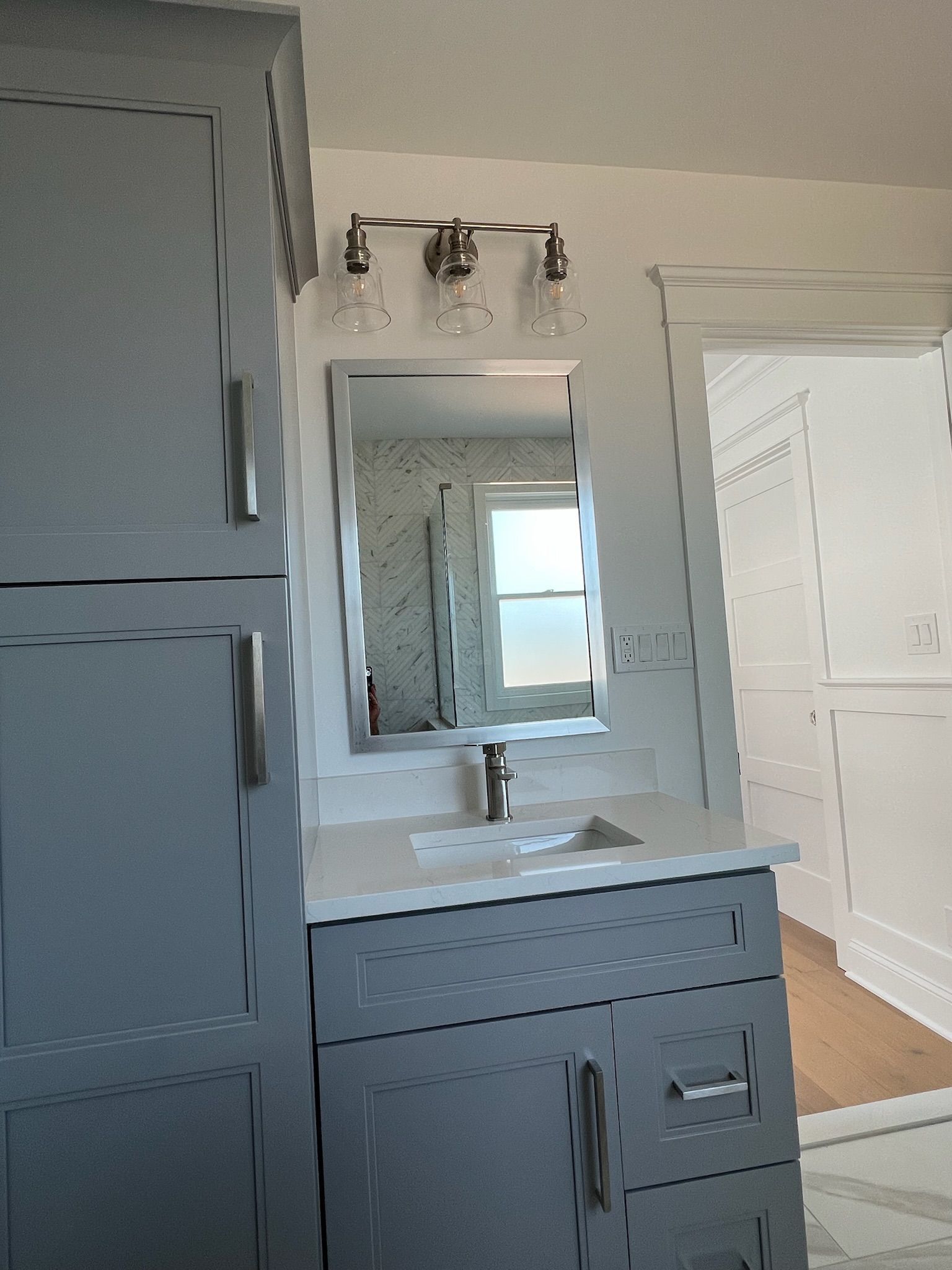 A gray bathroom vanity with a white countertop, silver faucet, rectangular mirror, and a three-light fixture above.