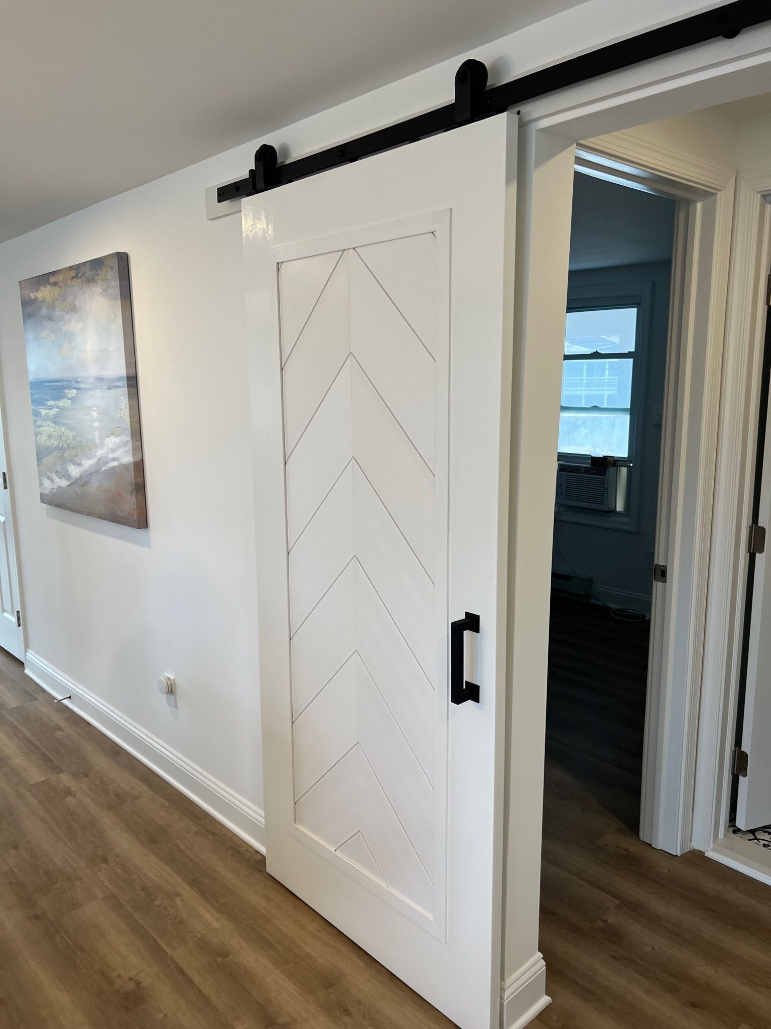 A white chevron-patterned sliding barn door with black hardware installed in a hallway with light wood-look flooring.