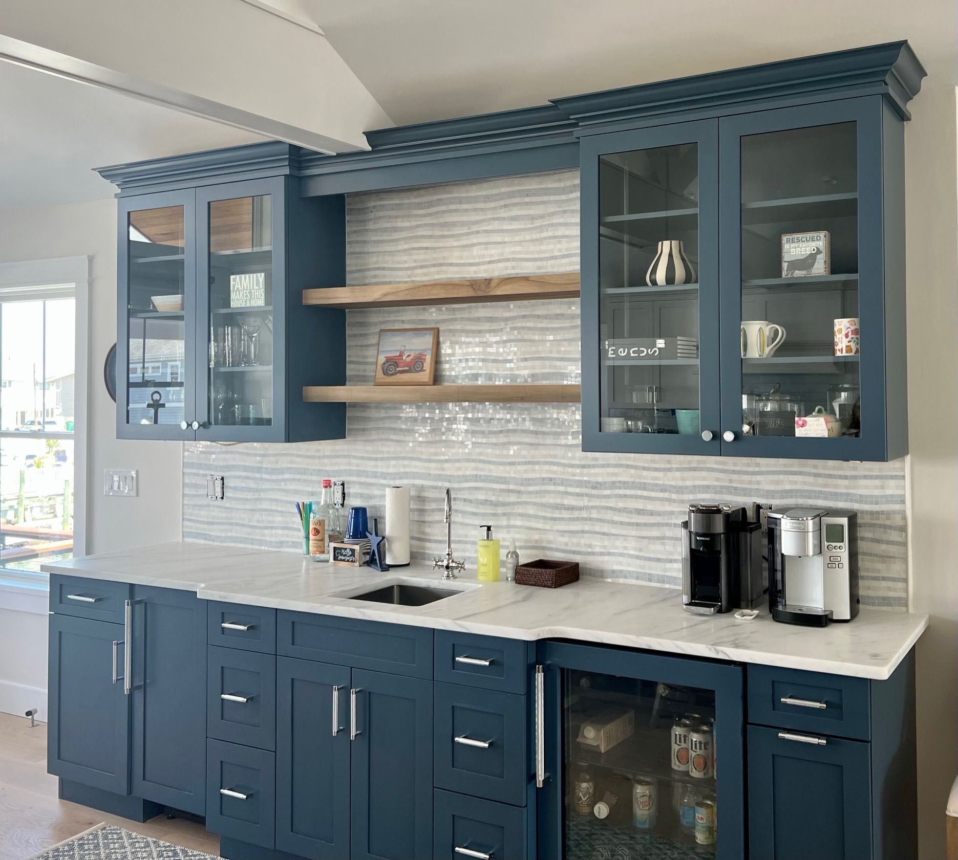 A kitchen wet bar with navy blue cabinets, white countertops, two wooden floating shelves, and a tiled backsplash.