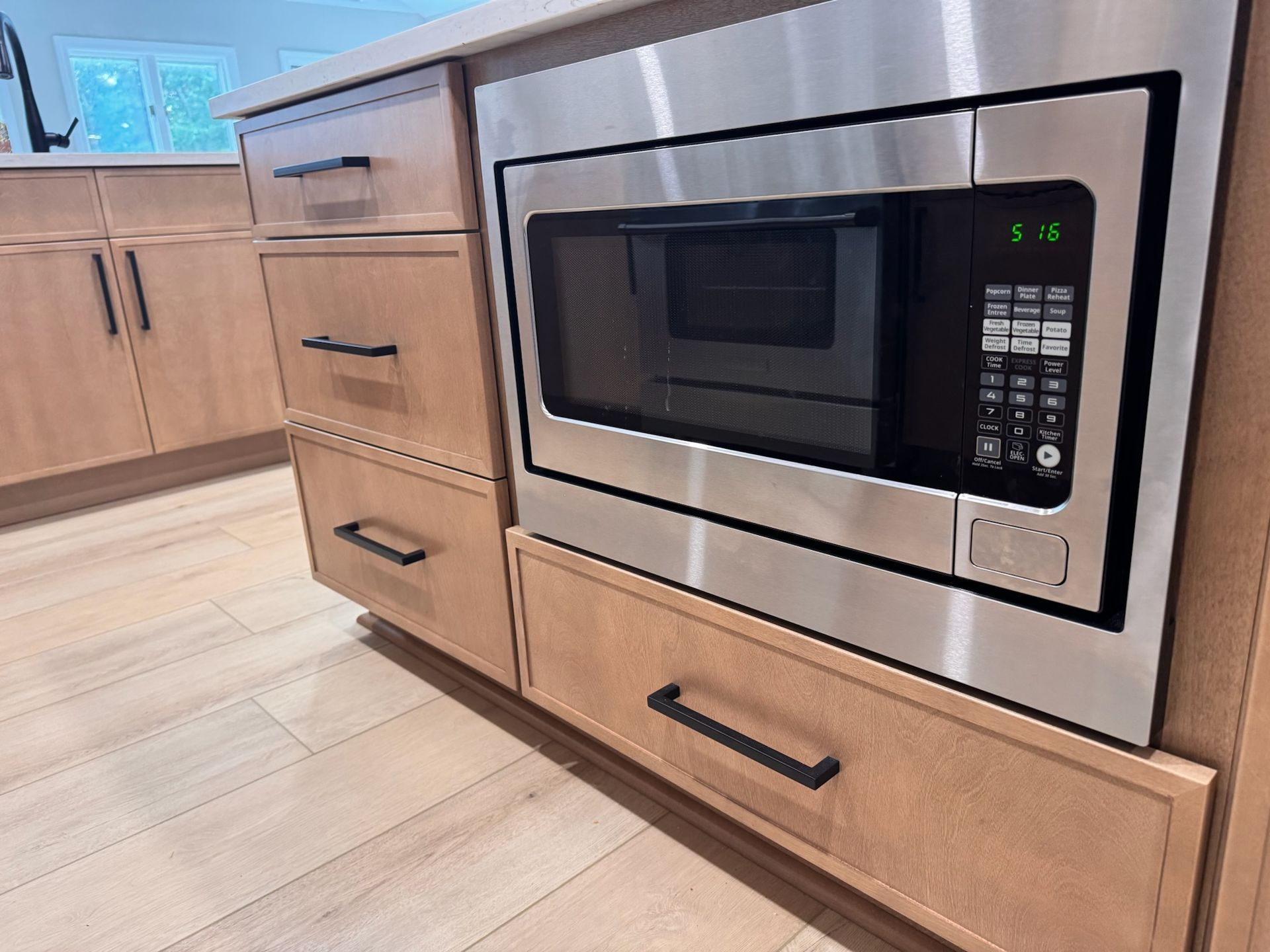 A built-in stainless steel microwave set into light wood kitchen cabinetry with black hardware and wood-look flooring.