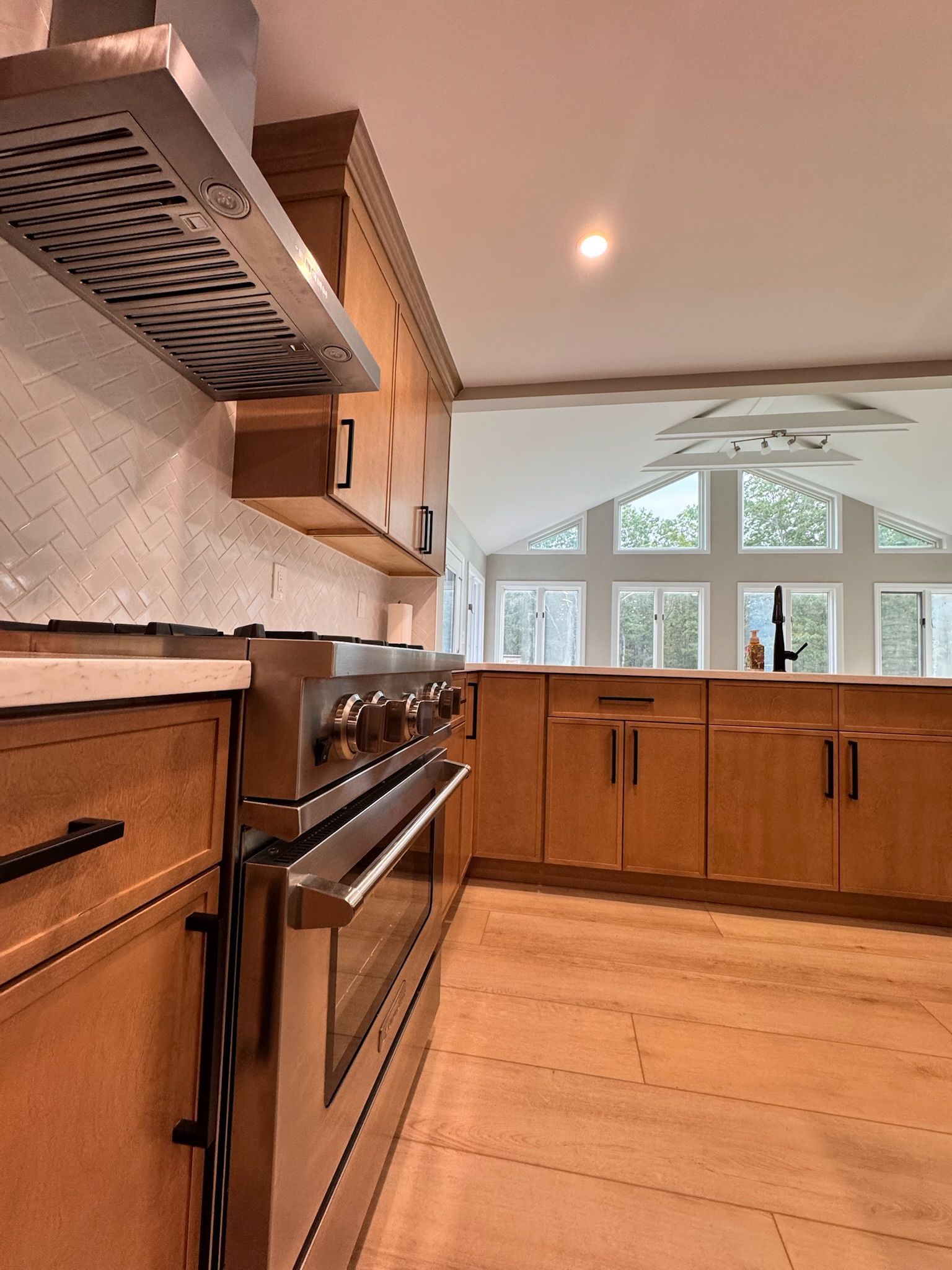 A modern kitchen with wooden cabinets, stainless steel range and vent hood, and large windows looking out at greenery.