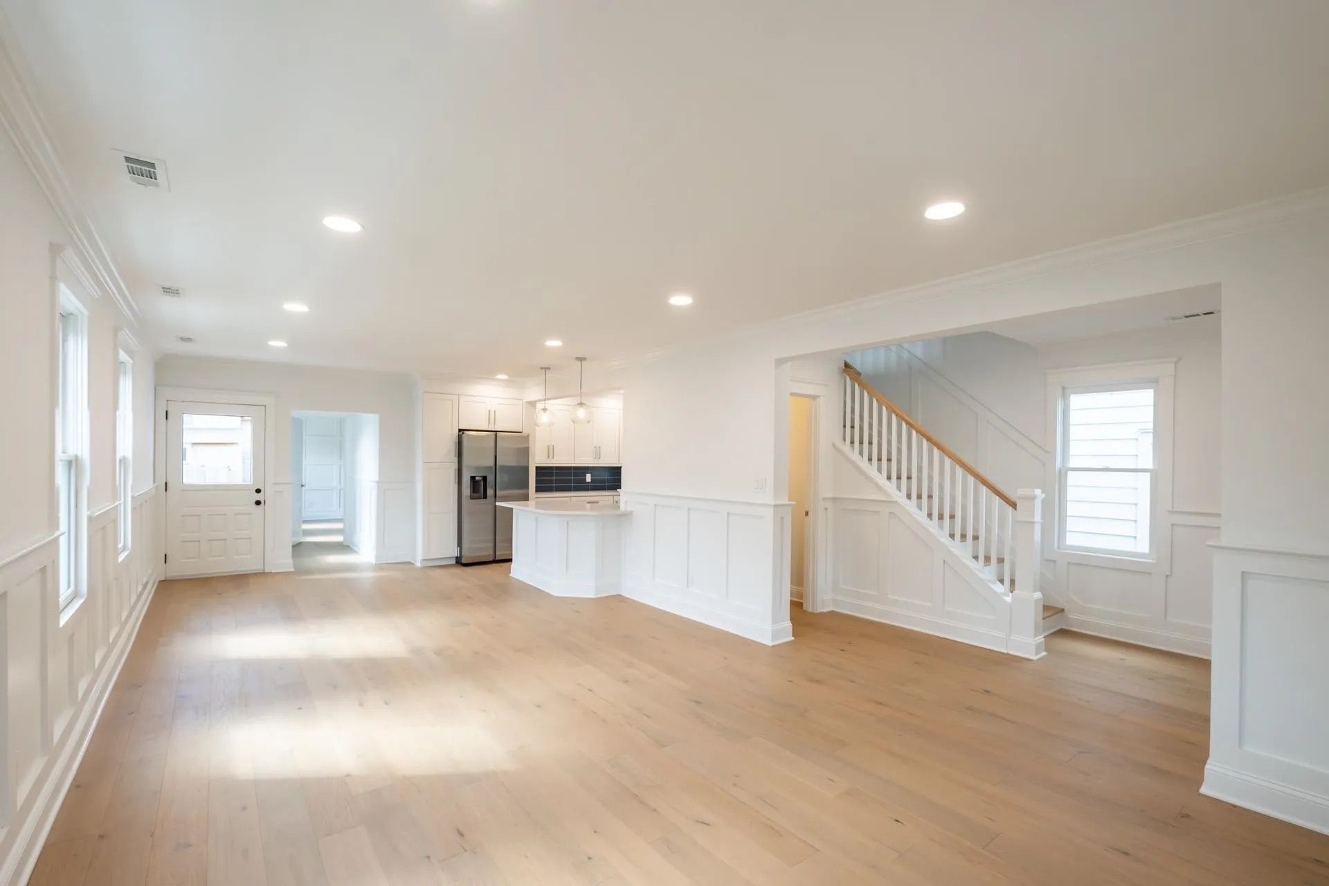 A spacious, light-filled room with white walls, wainscoting, light wood floors, a kitchen area, and a staircase.