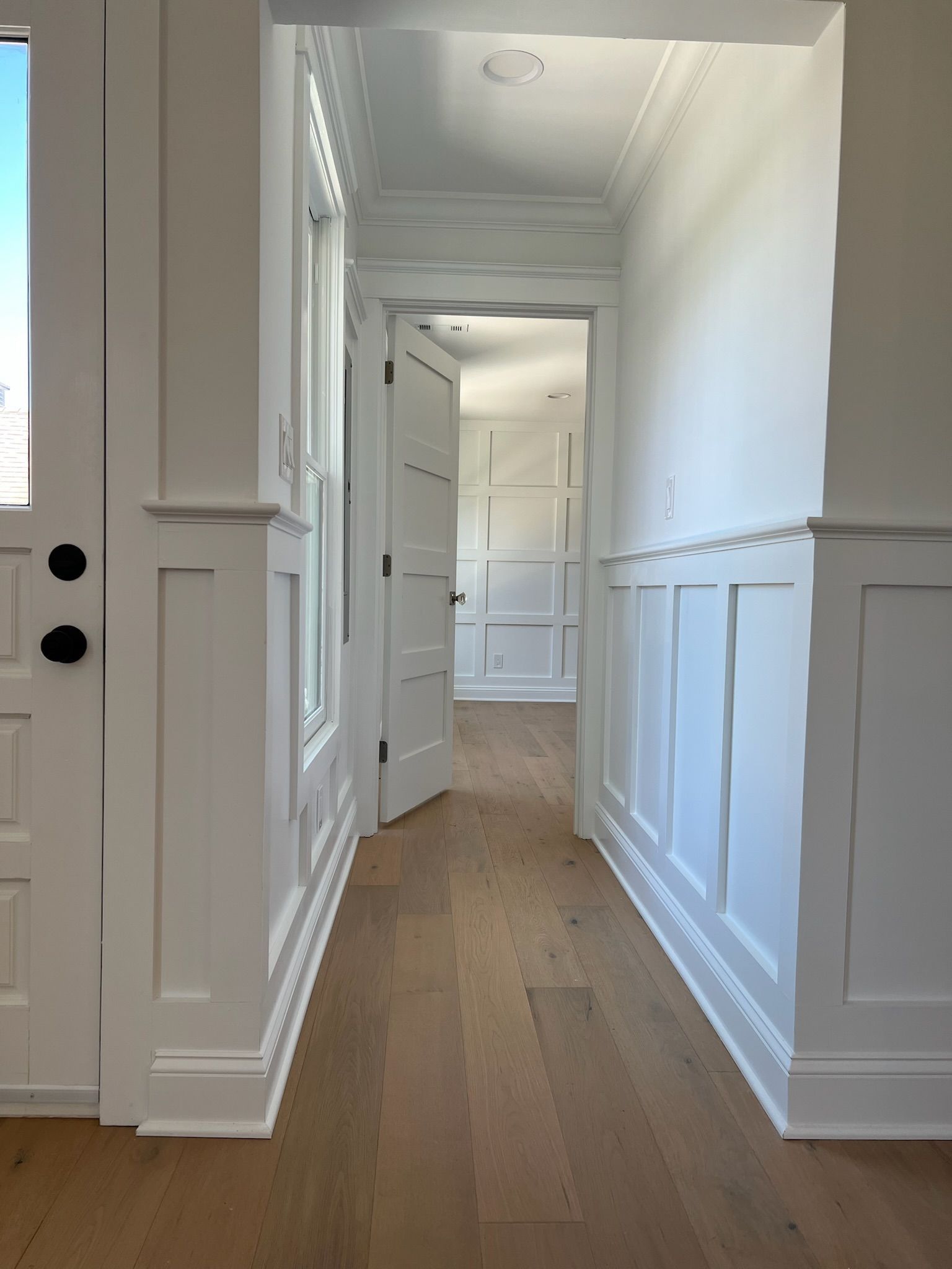 A bright hallway with light wood floors, white wainscoting on the walls, and a door opening into another paneled room.