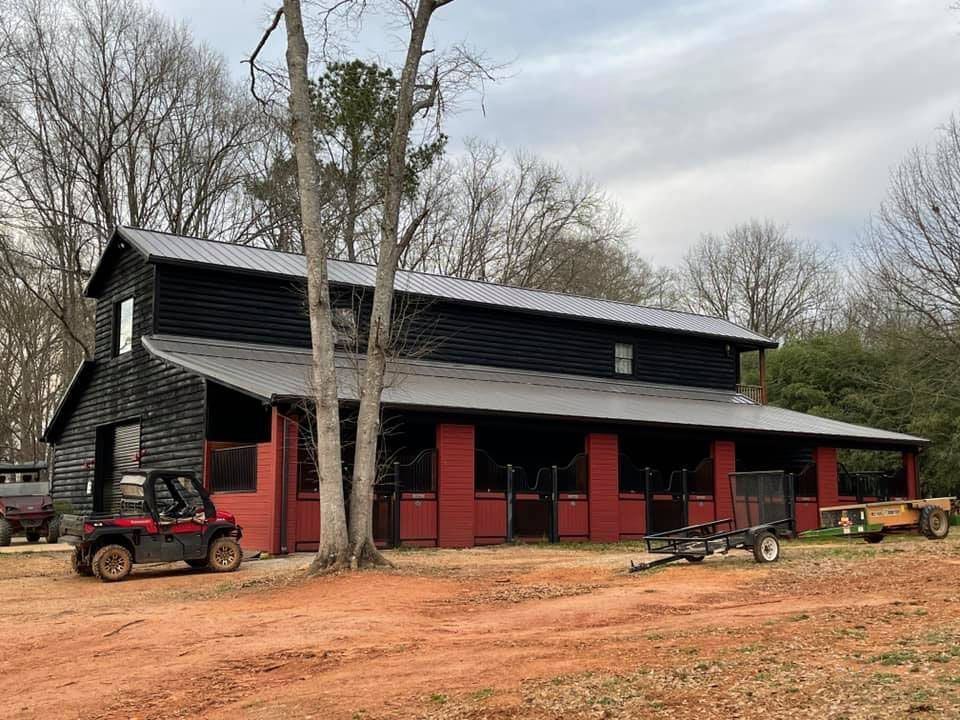 A red barn with a black roof is surrounded by trees and dirt