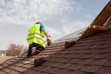 Worker in neon safety vest repairs shingles on a sloped roof under a cloudy sky