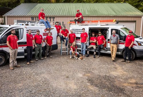 Group in red shirts posing with trucks and a dog outside a metal building
