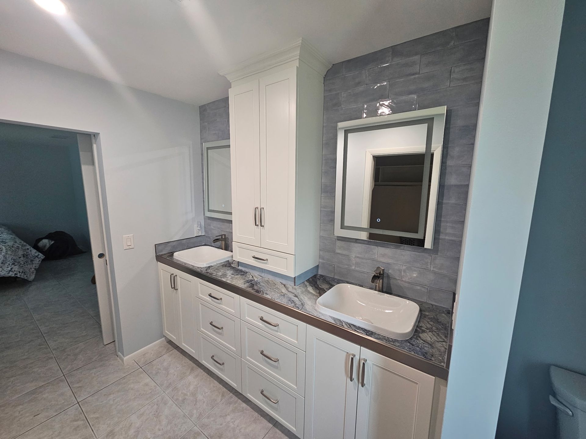 Bathroom with dual sinks, white cabinets, gray countertop, and a blue tiled wall. A tall cabinet and mirrors are also present.