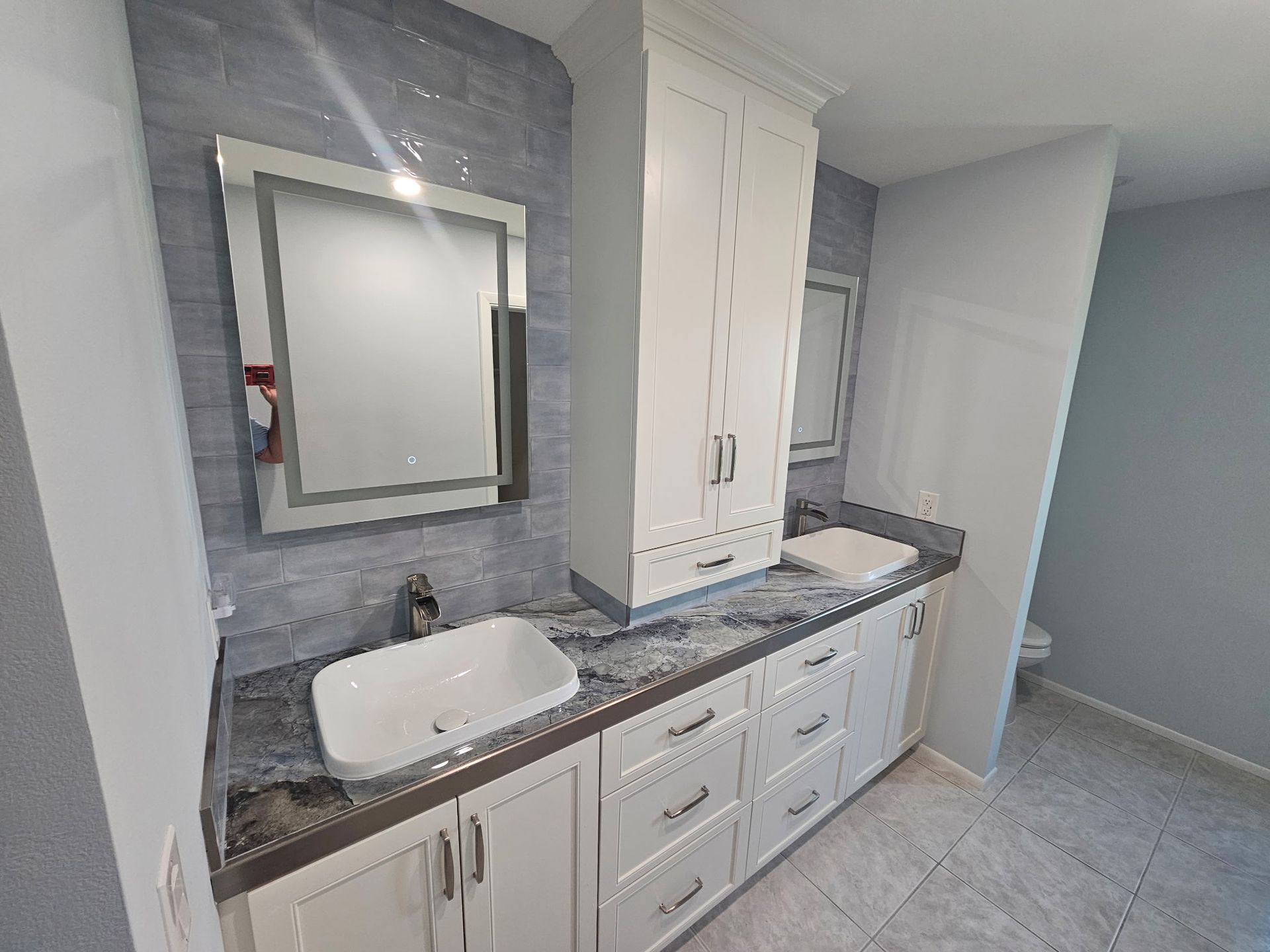 Bathroom with white cabinets, a blue tiled wall, and a large mirror above the sinks. The countertop is a mottled blue and gray.