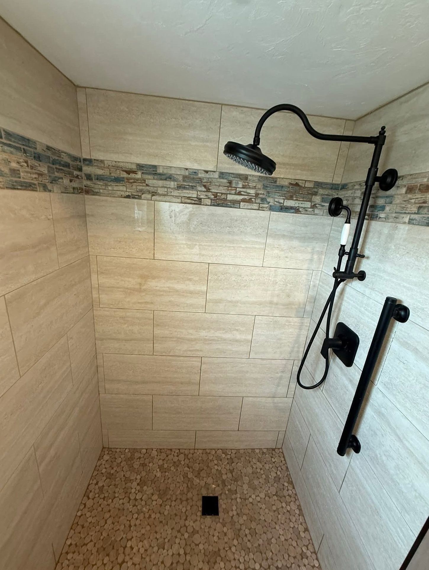 Shower with beige tiled walls, pebble floor, black fixtures, and a grab bar.
