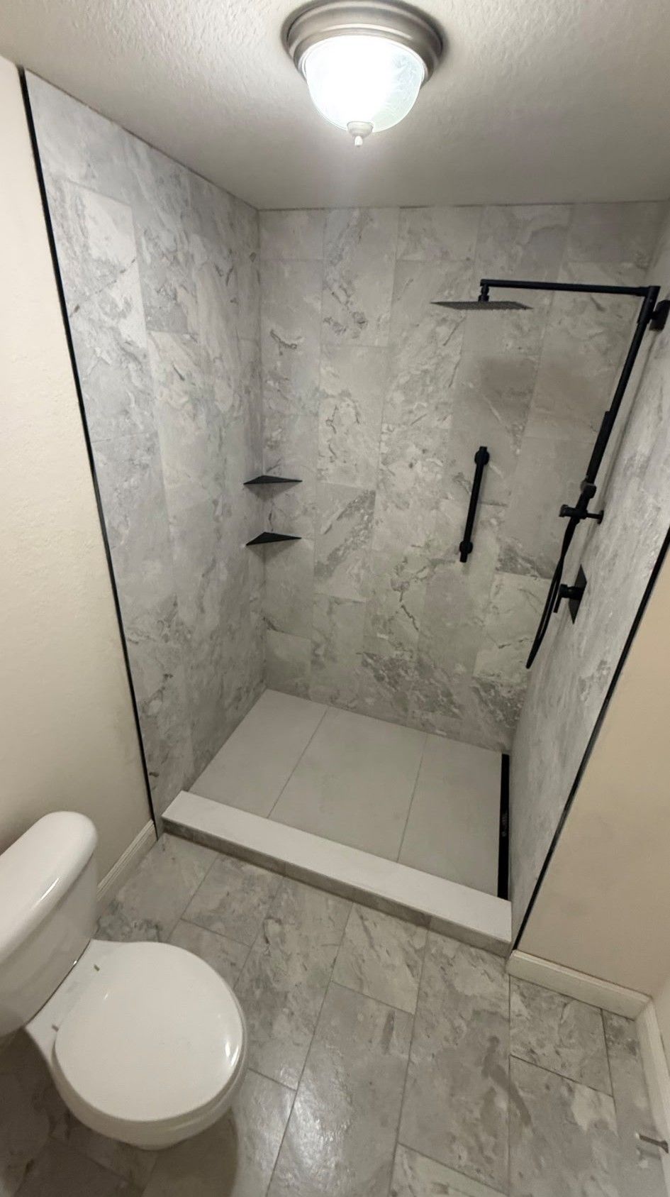 Bathroom with gray tiled shower and black fixtures, toilet in the foreground.