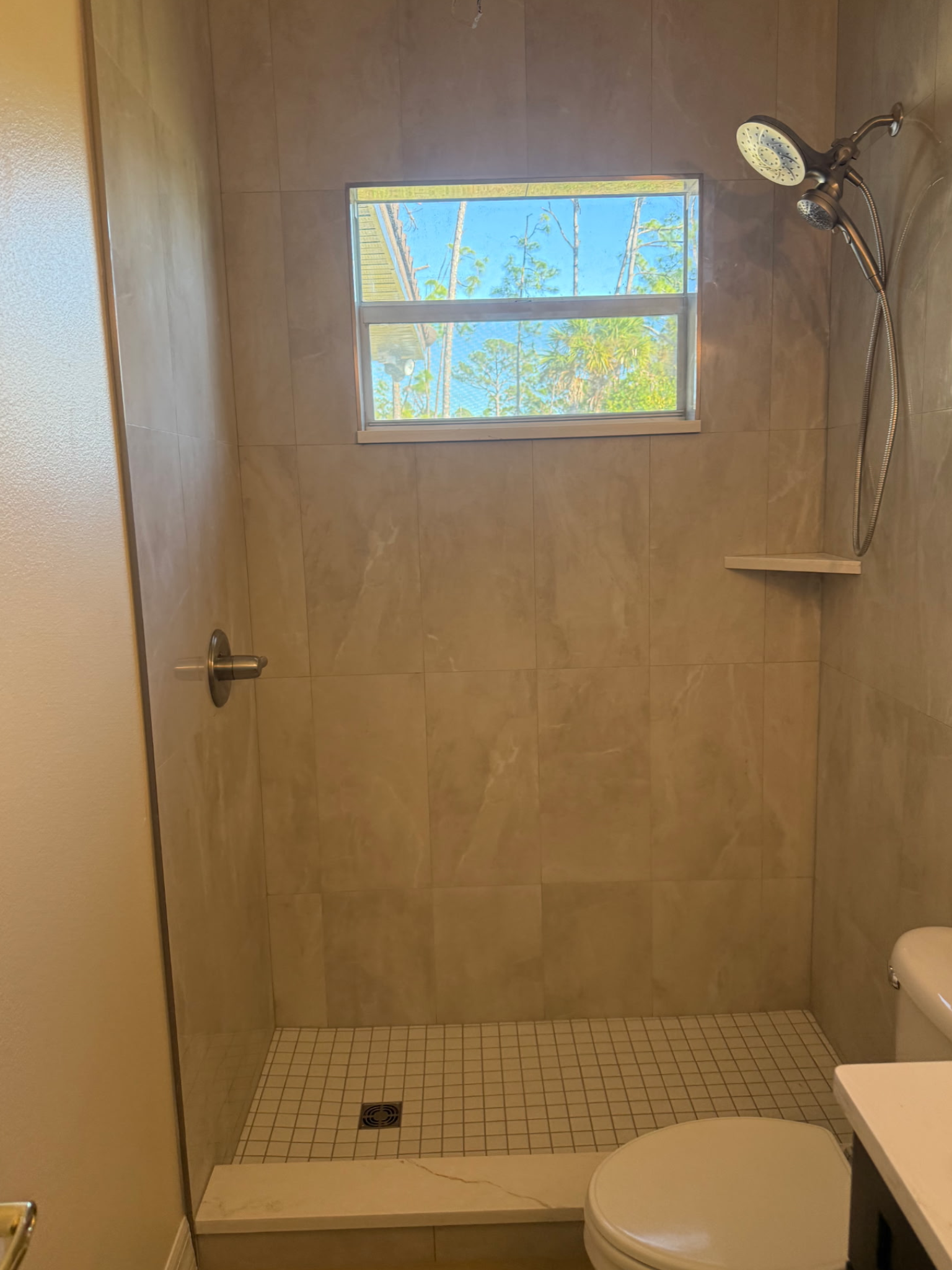 Shower stall with beige tile, small window, and chrome shower head and handle. Toilet in the foreground.