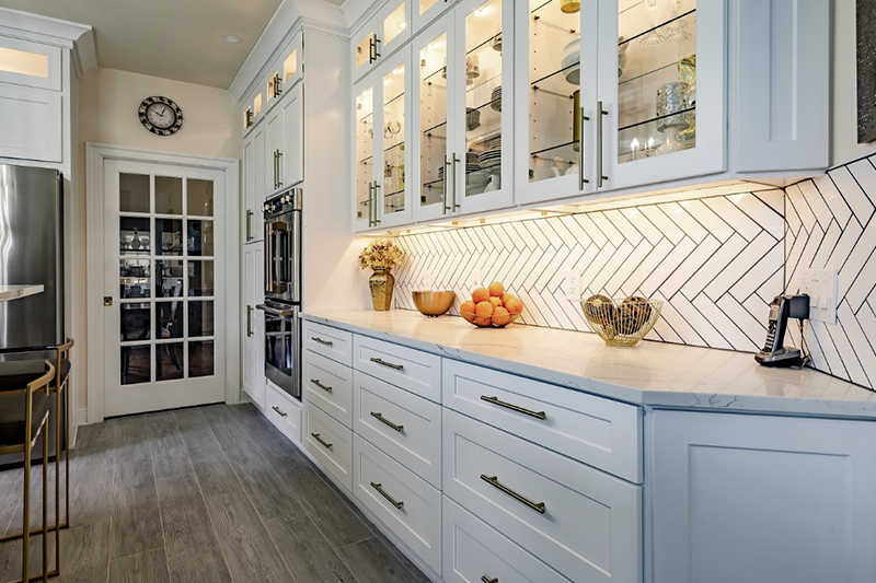 Bright modern kitchen with white cabinets, glass fronts, chevron backsplash, and stainless steel appliances.