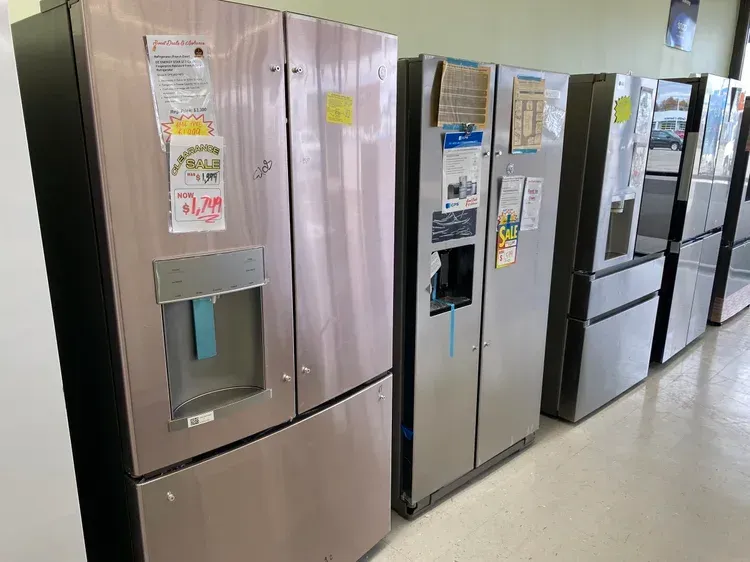 Row of stainless steel refrigerators in a store, some with water dispensers and various tags.