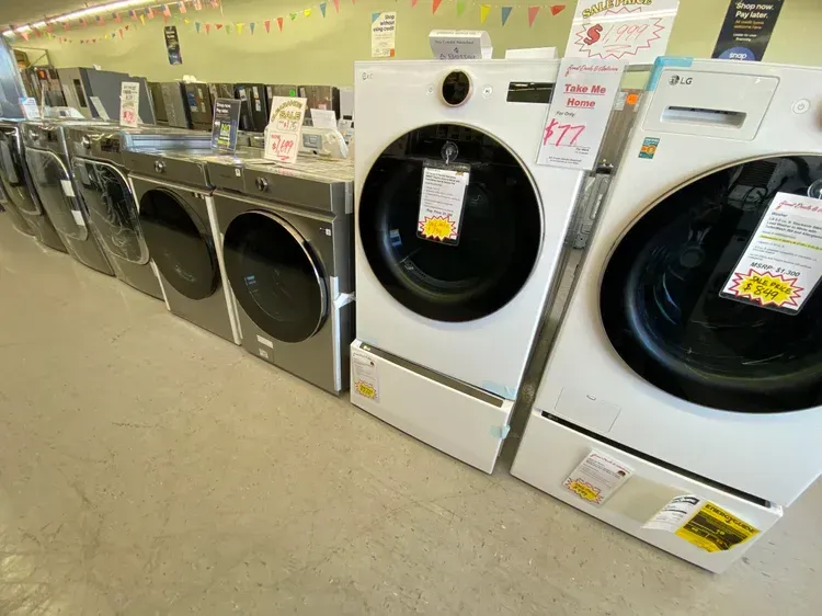 Row of washing machines in a store; white and grey, front-load models, price tags attached.