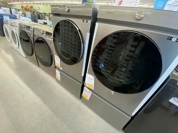 A row of Samsung washing machines and dryers in a store; various colors, shiny surfaces, glass doors.