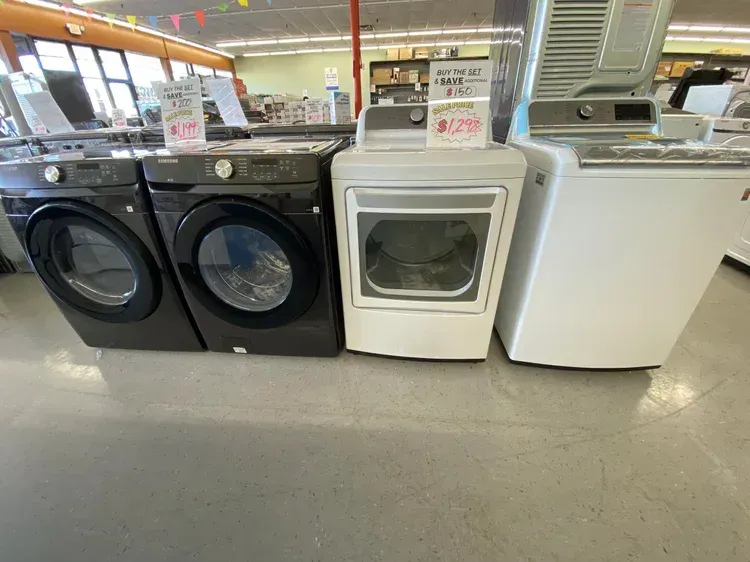 Washing machines and dryers for sale in a store. Black, white, and gray machines lined up on a floor.