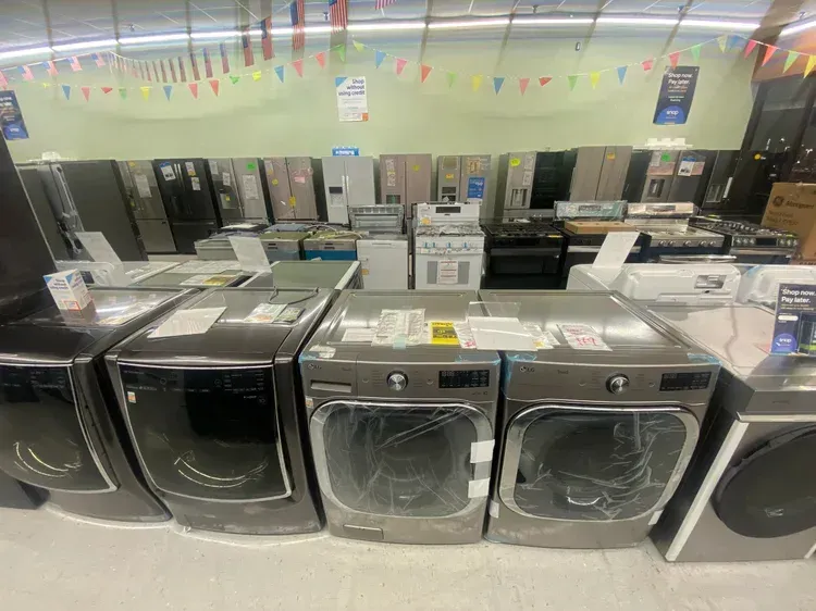 Washing machines and dryers on display in a store. Gray and black appliances with price tags sit in a row.