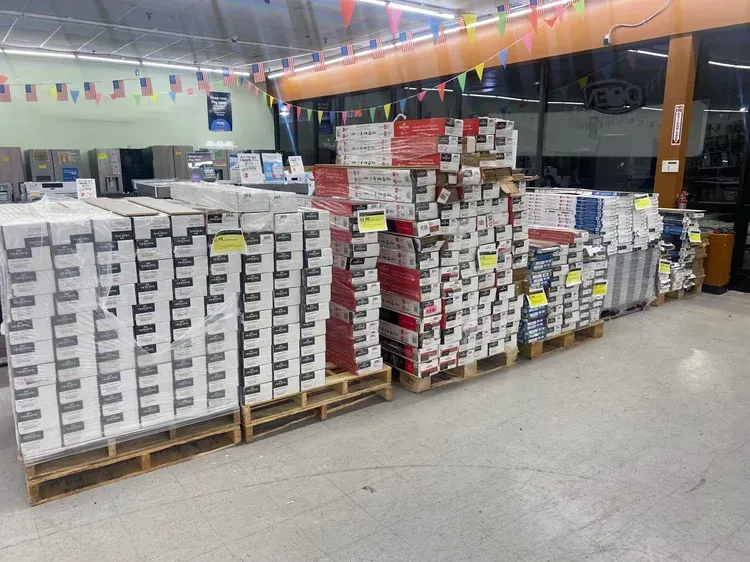 Pallets of boxes stacked in a store, possibly tile or flooring, under bright lights.