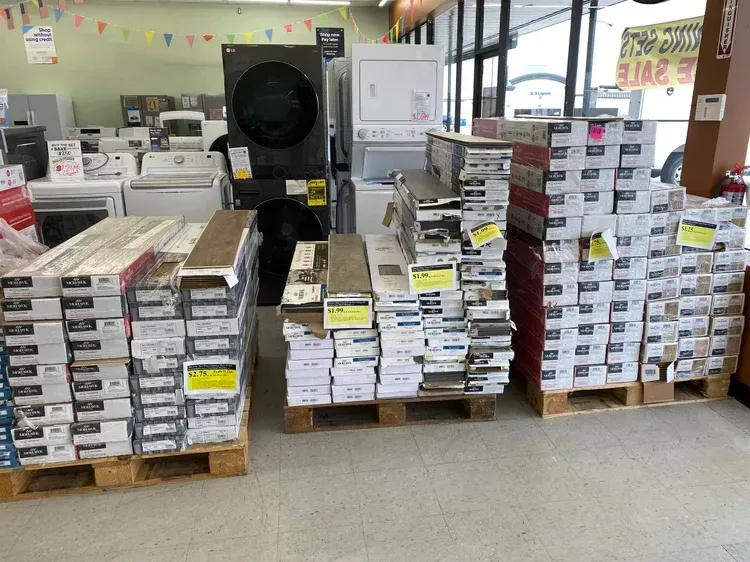 Pallets of flooring in a store, with appliances in the background and a sale sign.