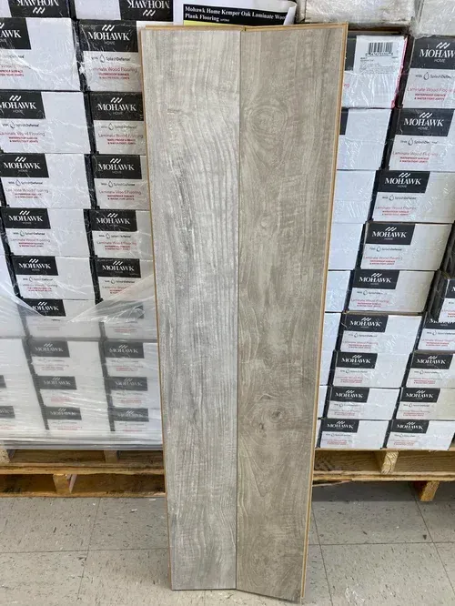 Wooden floorboard sample, light gray color, on a pallet with boxes of flooring in the background.
