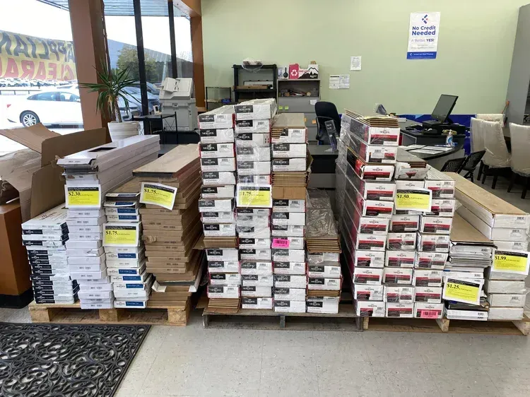 Stacks of flooring boxes on pallets inside a store. Sunlight streams through the window.