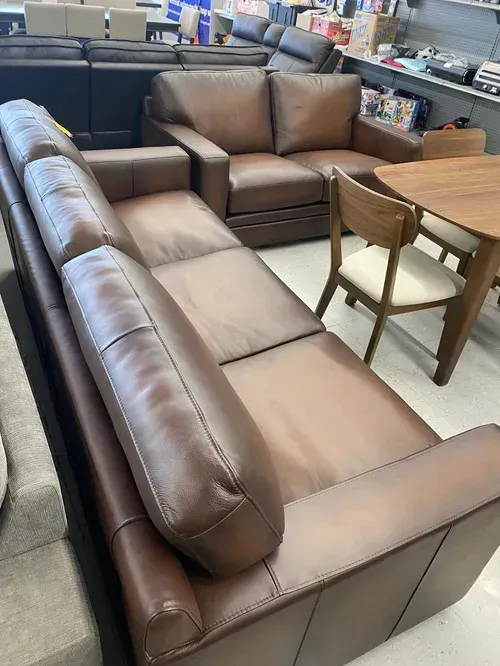 Brown leather sectional sofa and loveseat in a furniture store. A wooden dining table and chair are visible.