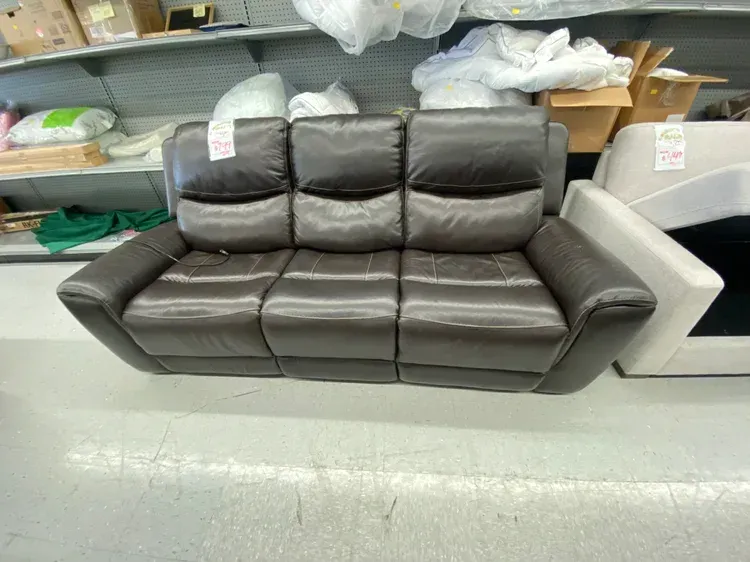 A brown leather reclining sofa in a store, with a white and gray floor and shelves in the background.