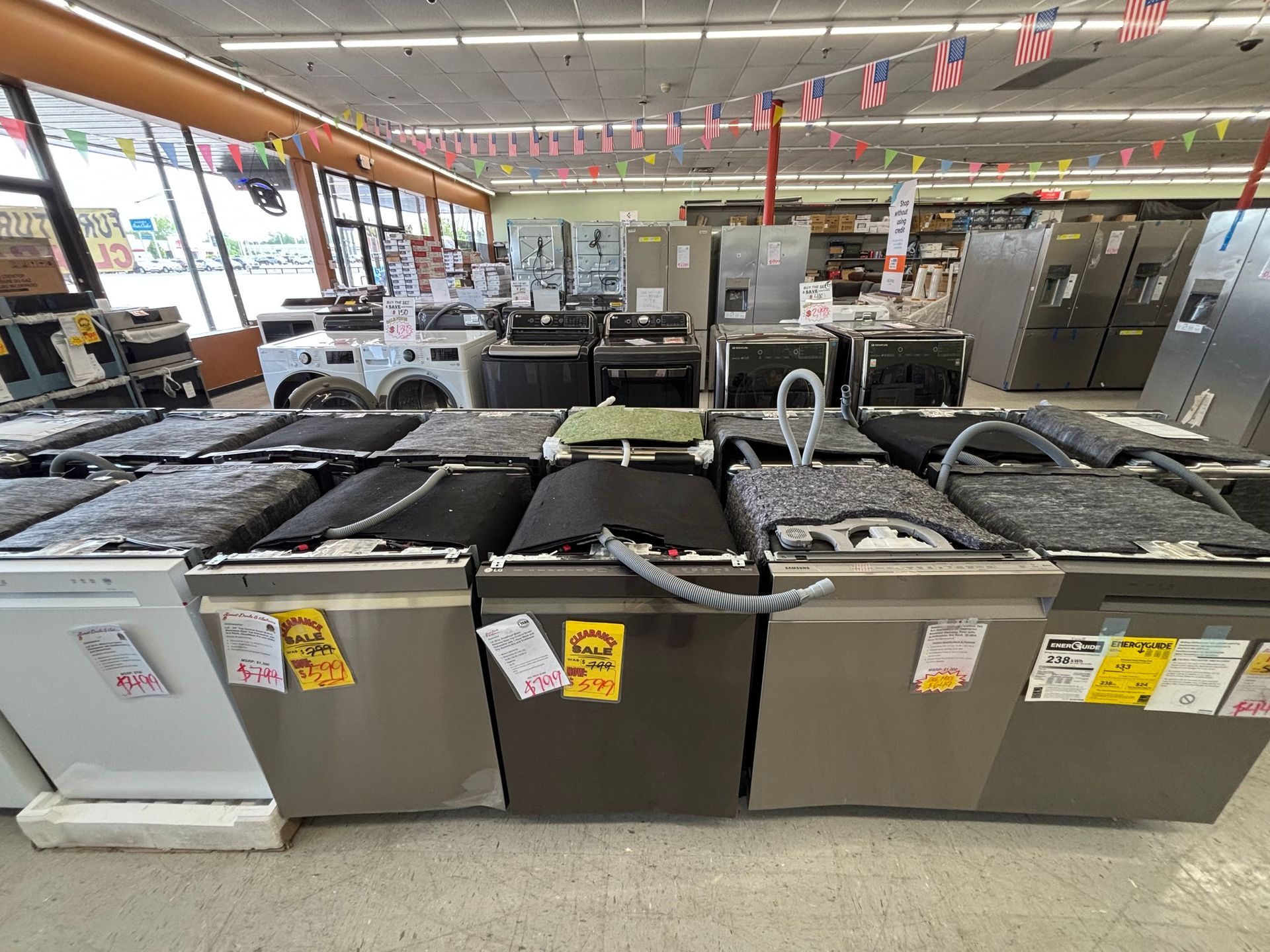 Rows of dishwashers on display in an appliance store. Refrigerators and washing machines are in the background.