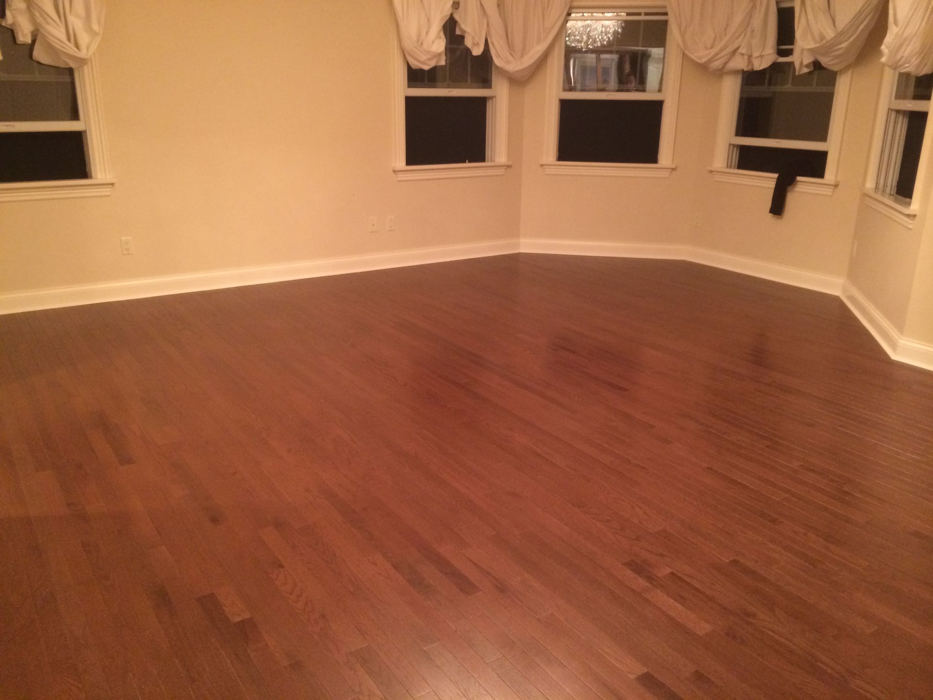 Empty room with hardwood floors and windows dressed with white curtains.