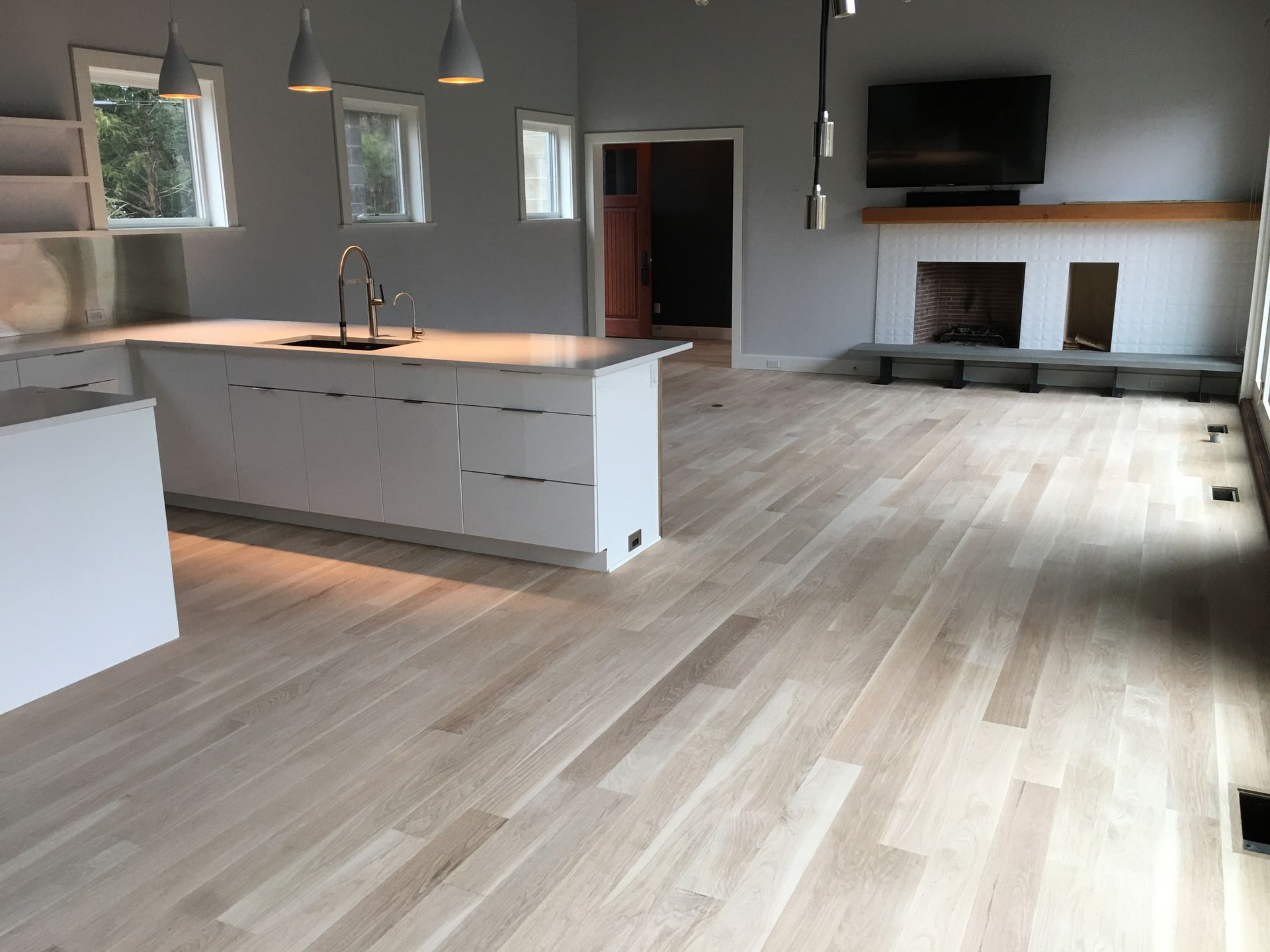 Empty modern kitchen and living area with light wood floors, island pendant lights, and a wall-mounted TV above a fireplace.
