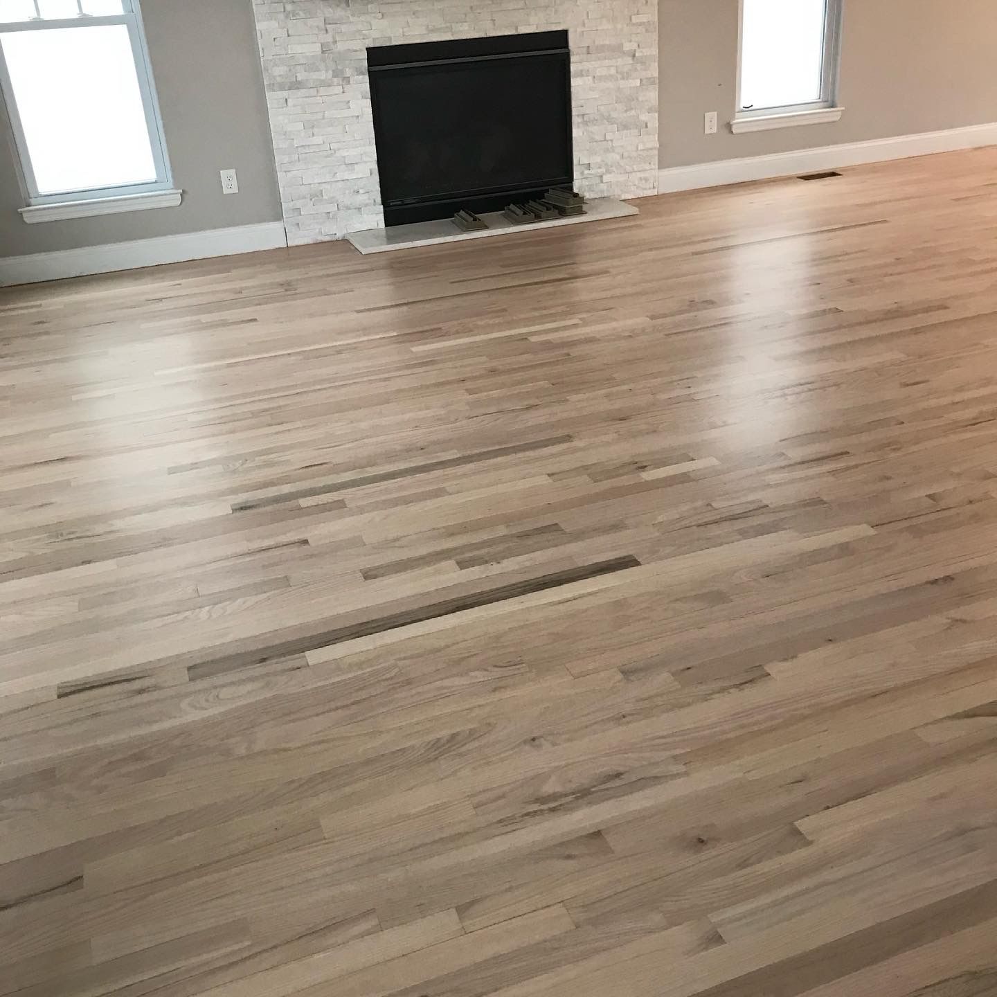 Spacious empty living room with light wood floors, a tiled fireplace, and two windows with blinds