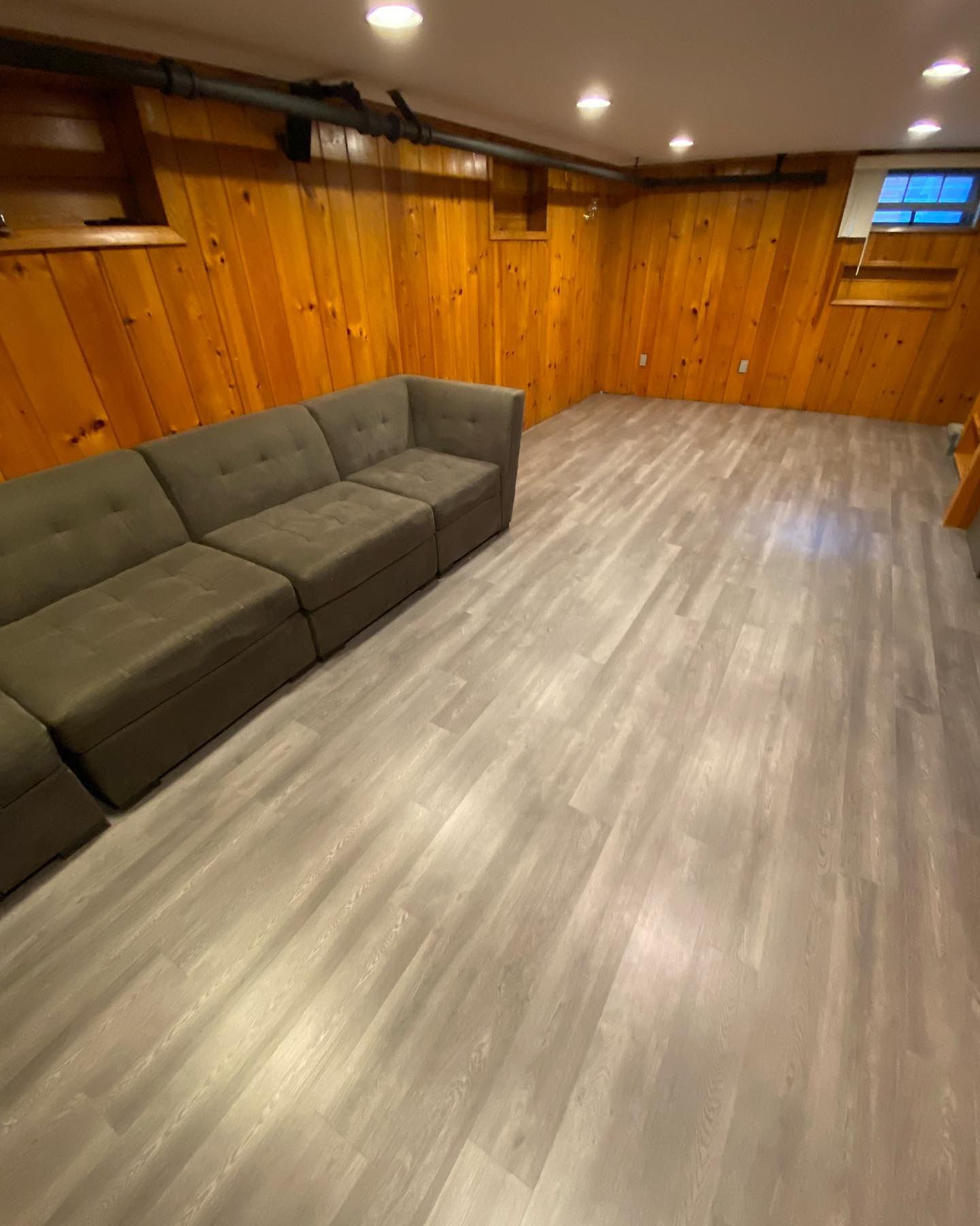 Empty basement with wood-paneled walls, gray sectional sofa, and light wood flooring under recessed lights