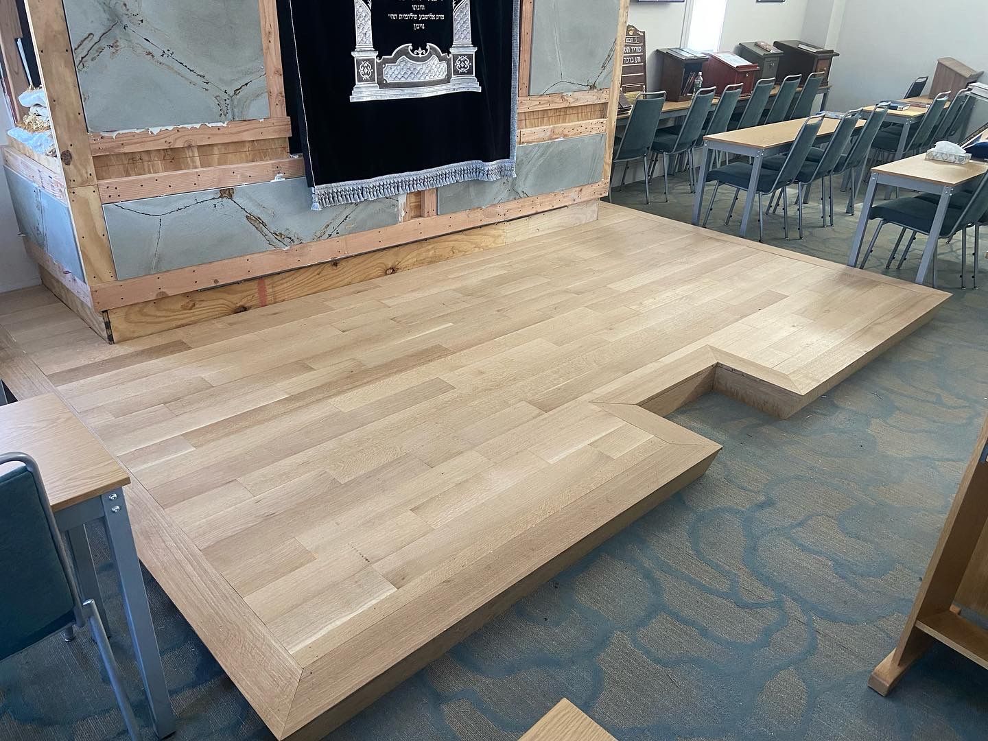 Wooden stage platform in a room, with a raised section and recessed cutout near chairs and a wall display.