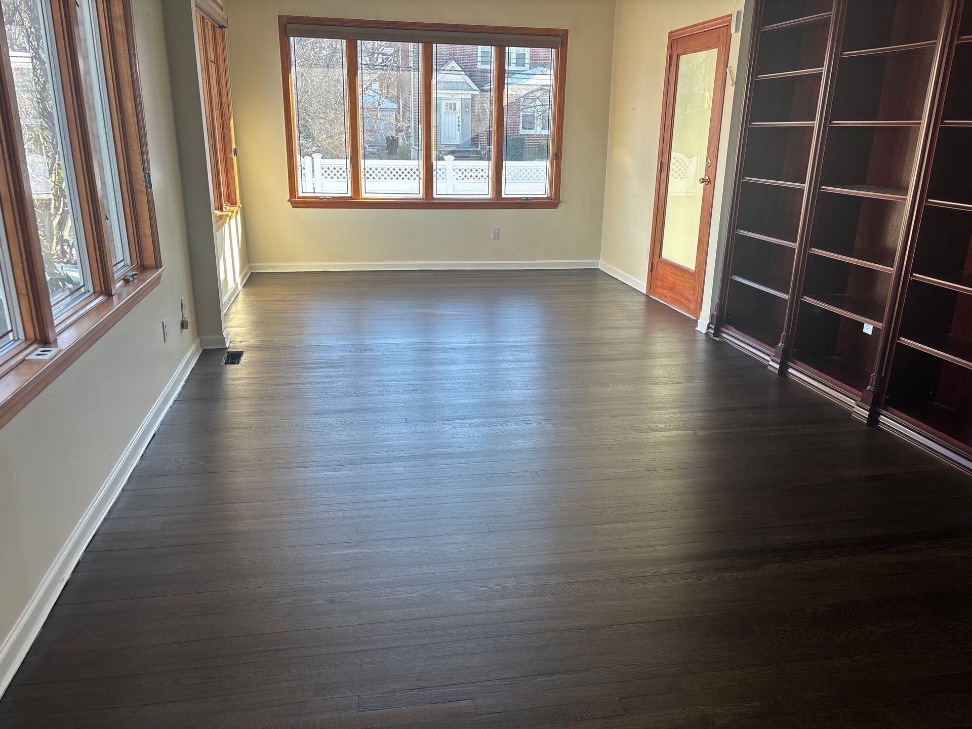Empty room with dark wood floors, large windows, and built-in shelving along one wall