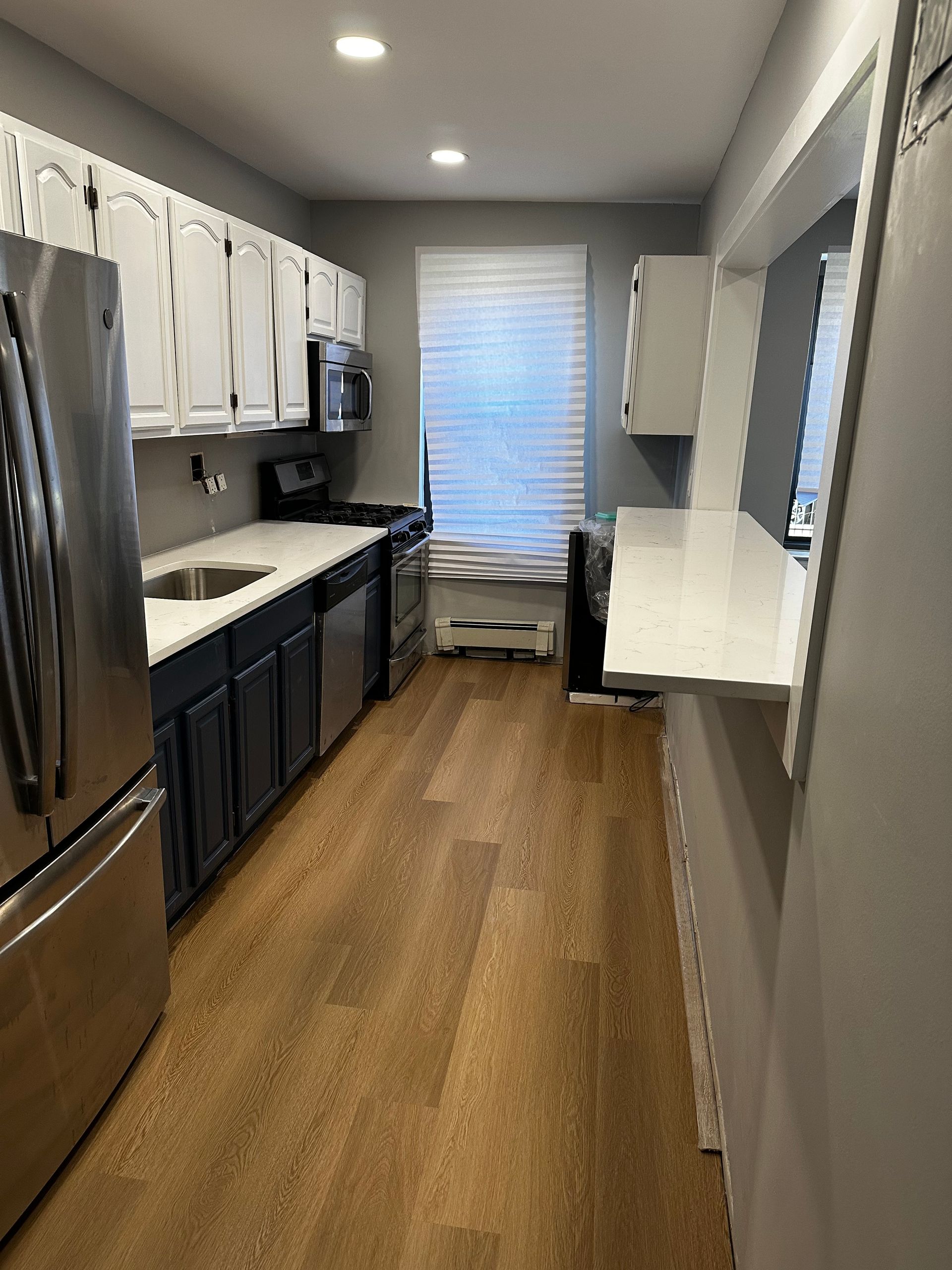 Narrow galley kitchen with dark cabinets, stainless fridge, white countertops, and window at the end.