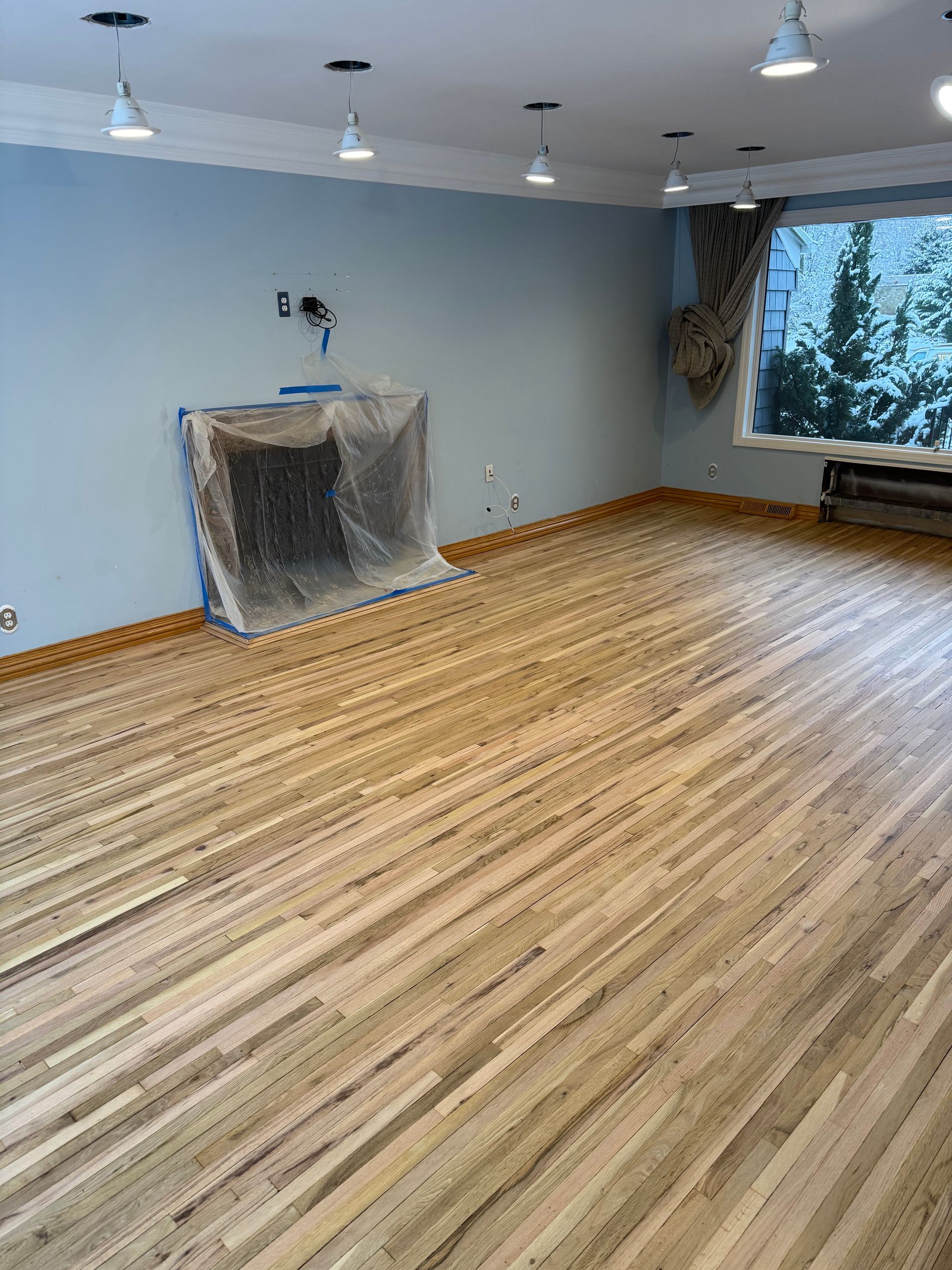 Empty room with light blue walls, wood floor, ceiling lights, and a window by a covered fireplace.