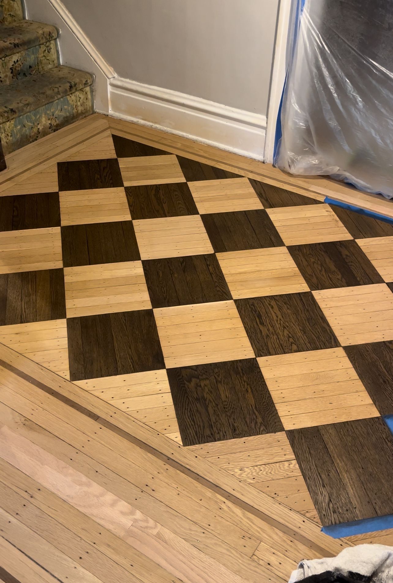 Partially installed wood floor with a checkerboard inlay beside a staircase and plastic-covered wall