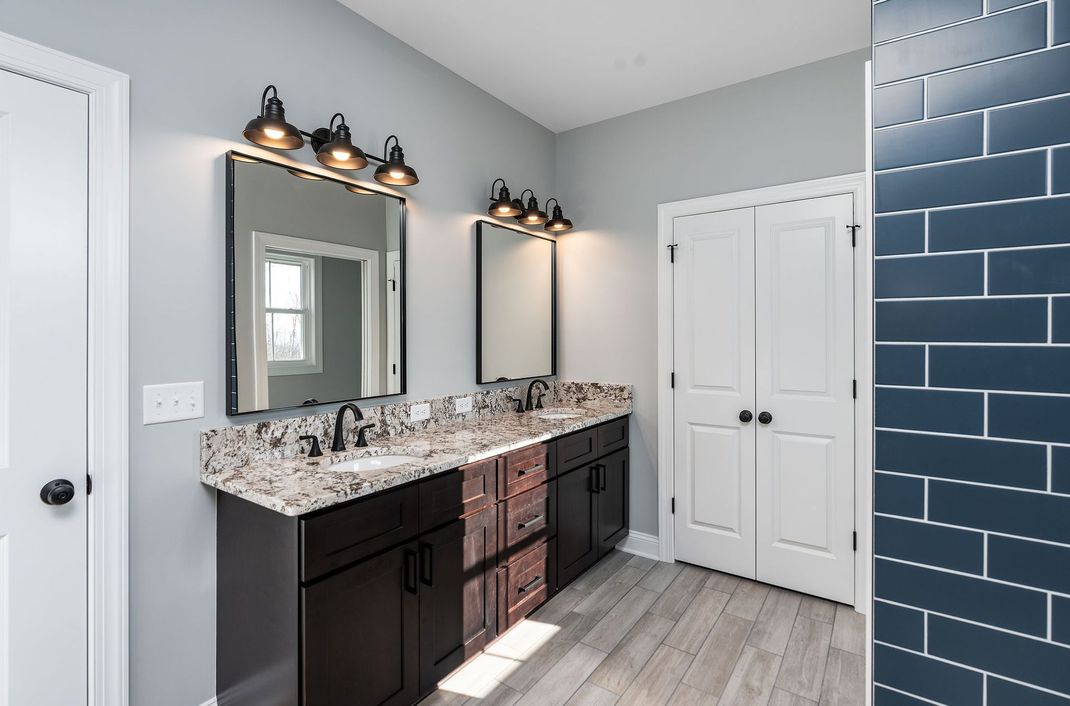 Bathroom with double sinks, dark cabinets, granite countertop, blue tile shower wall, and gray walls.