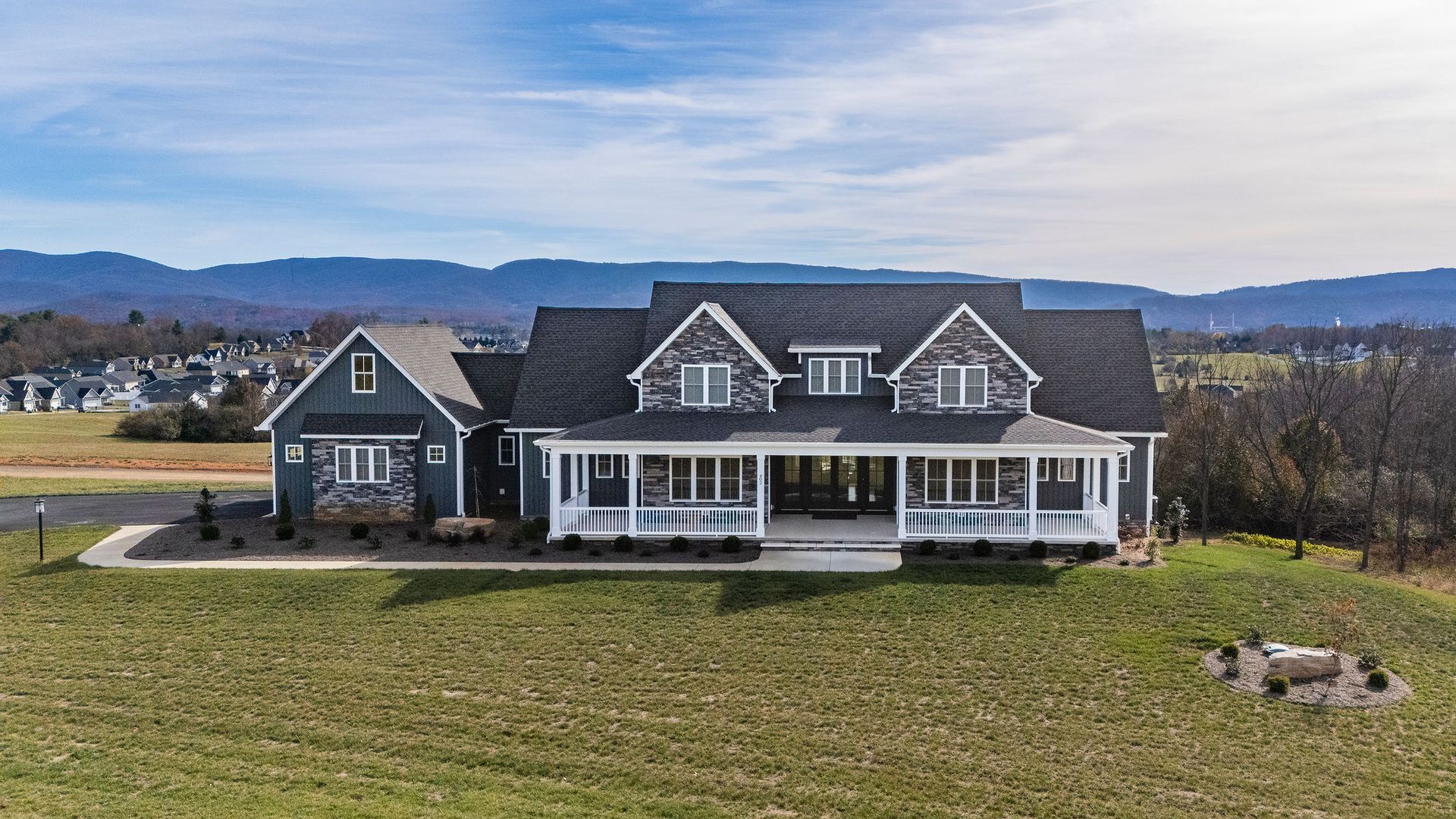 Blue-sided house with stone accents, white porch, and dark roof sits on a grassy hill; mountains in the background.