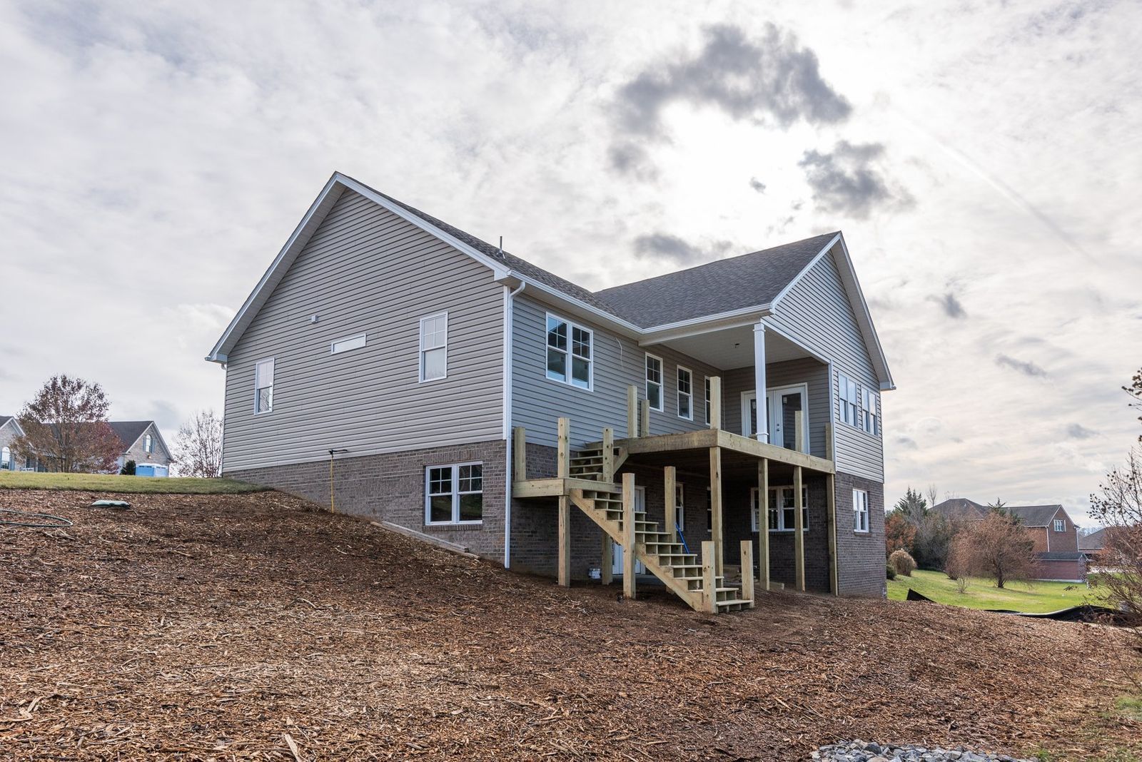 Two-story house with gray siding, brick foundation, wooden deck, and a cloudy sky.