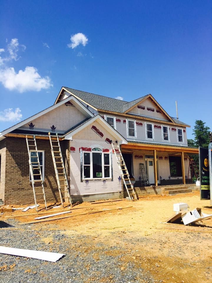 House under construction; exterior with ladders, unfinished siding, and brown brick. Bright blue sky.