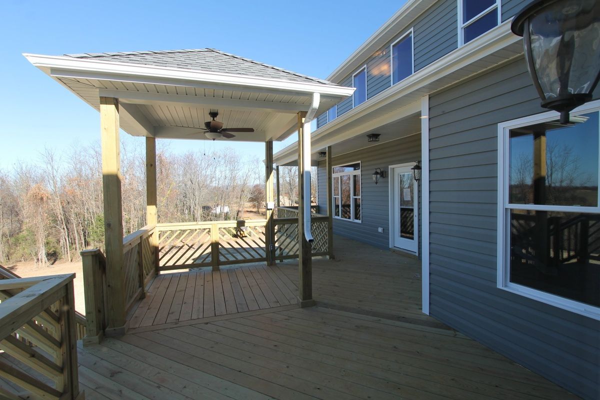 Wooden deck with gazebo, attached to a gray house, overlooking a wooded area.