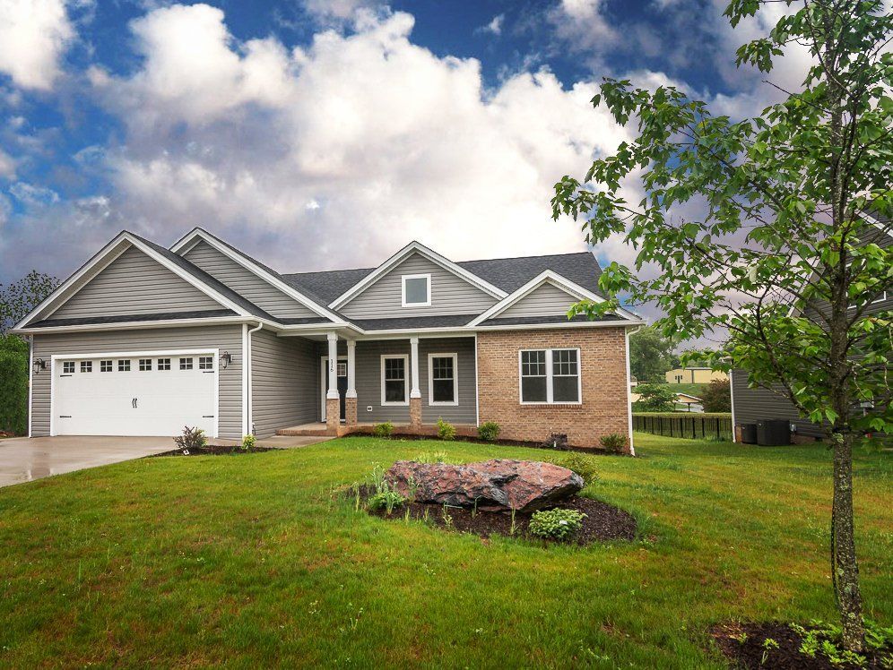 Suburban house with gray siding, brick accents, and a white garage door, set on a green lawn under a cloudy sky.