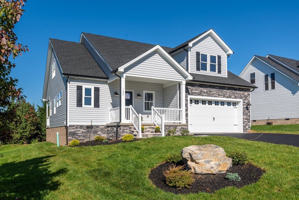 White and gray house with a stone accent and a garage, on a grassy hill under a blue sky.