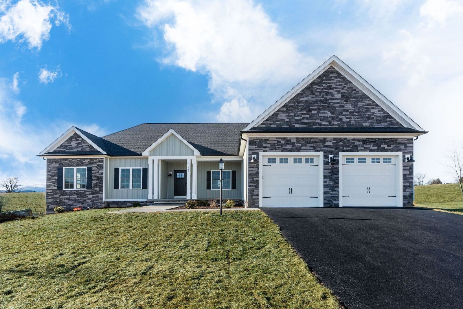Ranch-style house with gray and stone exterior, two-car garage, and asphalt driveway on a grassy hill under a blue sky.