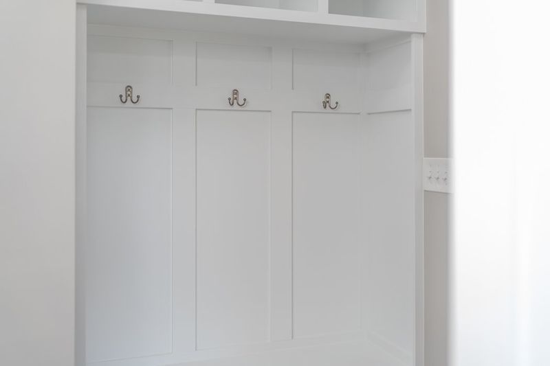 White built-in coat rack with three hooks, flanked by vertical panels and overhead shelf.