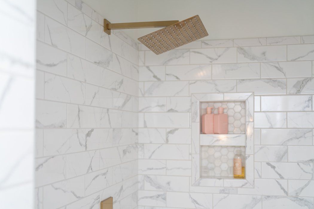 Shower with white marble-look tiles, a gold showerhead, and a recessed shelf holding toiletries.