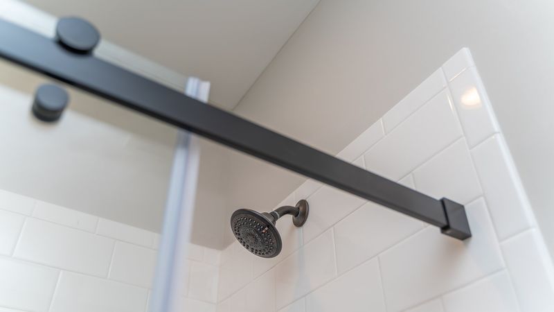 Black shower head and sliding door track against white tiled wall.