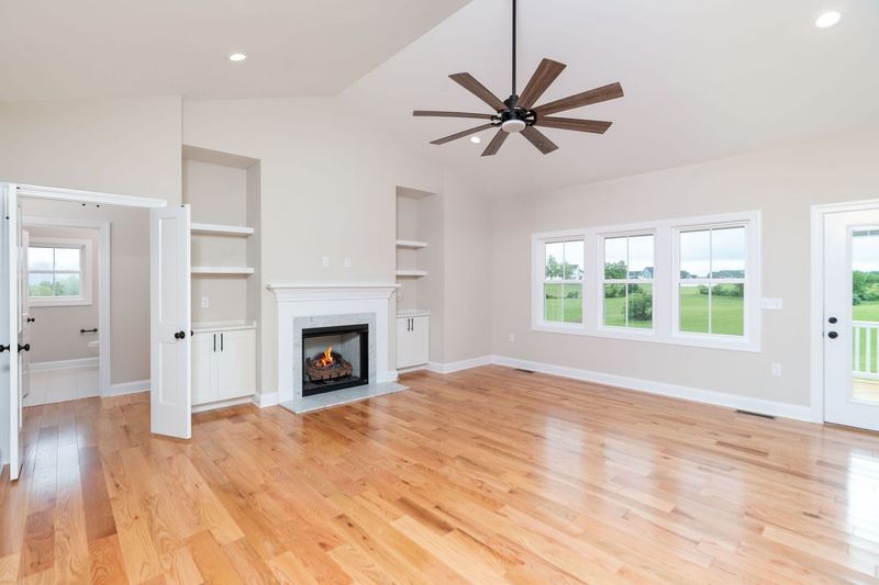 Spacious living room with hardwood floors, fireplace, built-in shelves, and large windows overlooking a green field.